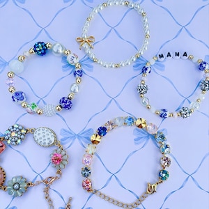 Puede incluir: Una variedad de pulseras de cuentas en varios estilos. Una pulsera presenta la palabra "MAMA" en cuentas blancas. Otras pulseras incluyen perlas, gemas de colores y detalles florales. El fondo es un patrón azul claro con lazos.