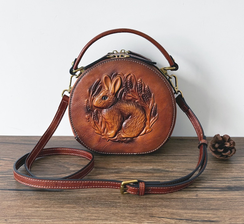 May include: A round, brown leather handbag with a detailed embossed design of a rabbit surrounded by wheat. The bag has a top handle and a long, adjustable shoulder strap. The hardware is gold-colored.