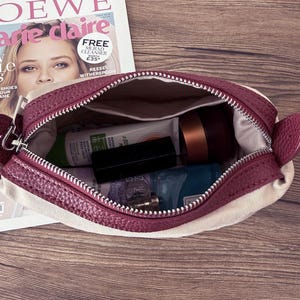 Puede incluir: Bolsa de cosm&eacute;ticos burdeos y crema abierta, con cierre de cremallera. Contiene varios productos de belleza, como una crema facial, un l&aacute;piz labial y un frasco peque&ntilde;o. Una revista est&aacute; en el fondo.