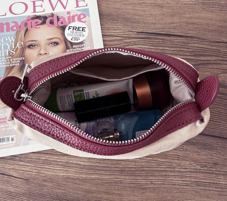 Puede incluir: Bolsa de cosm&eacute;ticos burdeos y crema abierta, con cierre de cremallera. Contiene varios productos de belleza, como una crema facial, un l&aacute;piz labial y un frasco peque&ntilde;o. Una revista est&aacute; en el fondo.