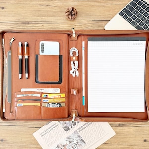 May include: A brown leather portfolio with a zipper closure, a phone pocket, card slots, and a memo pad. The portfolio is open and has a white memo pad with the word "MEMO" printed on it.