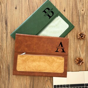 Puede incluir: Dos fundas de documentos de cuero personalizadas, una marrón y otra verde, se muestran sobre una superficie de madera. La funda marrón presenta un bolsillo con cremallera y la letra "A" con "Your name" debajo. La funda verde tiene la letra "B" y "Your name".
