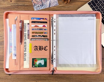 May include: A peach-colored zippered portfolio organizer featuring a metal three-ring binder, multiple card slots, a pen holder with a silver and red pen, a vertical document pocket, and a memo pad. The interior includes the text ABC.