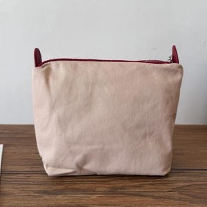 Puede incluir: Una bolsa de lona color rubor con cierre de cremallera y detalles de cuero burdeos. La bolsa tiene forma rectangular y un dise&ntilde;o sencillo, adecuada para transportar objetos peque&ntilde;os. La cremallera es plateada y la bolsa est&aacute; sobre una superficie de madera.