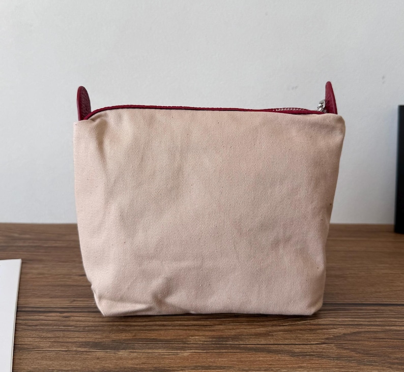 Puede incluir: Una bolsa de lona color rubor con cierre de cremallera y detalles de cuero burdeos. La bolsa tiene forma rectangular y un dise&ntilde;o sencillo, adecuada para transportar objetos peque&ntilde;os. La cremallera es plateada y la bolsa est&aacute; sobre una superficie de madera.