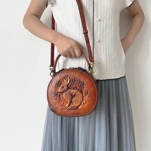 May include: A round, brown leather handbag with a detailed embossed design of a rabbit surrounded by wheat. The bag has a top handle and a detachable shoulder strap. The bag is approximately 8 inches in diameter.