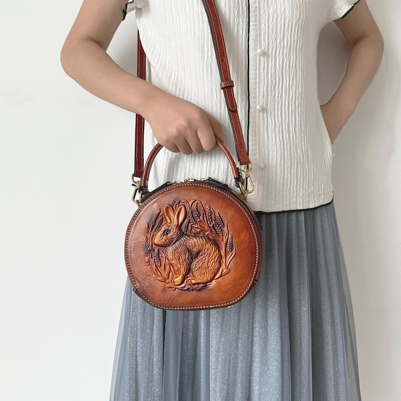 May include: A round, brown leather handbag with a detailed embossed design of a rabbit surrounded by wheat. The bag has a top handle and a detachable shoulder strap. The bag is approximately 8 inches in diameter.