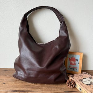 May include: A brown leather tote bag with a single strap. The bag is sitting on a wooden surface with a book and a picture frame.