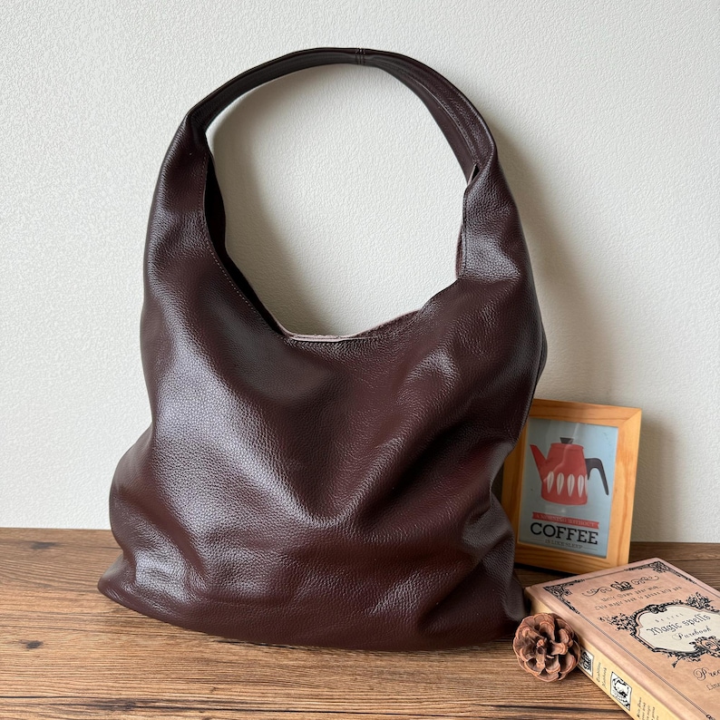 May include: A brown leather tote bag with a single strap. The bag is sitting on a wooden surface with a book and a picture frame.
