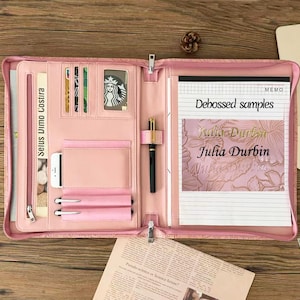 May include: A light pink zippered portfolio organizer featuring multiple interior card slots, a clear ID window, a vertical document pocket, two pen loops, and a central pen holder. A notepad with the text Debossed samples and Julia Durbin is included.