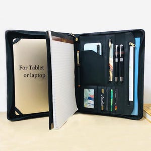May include: Black leather portfolio with multiple compartments, including slots for cards, pens, and a phone. The portfolio is open, revealing a notepad and a section labeled "For Tablet or laptop."