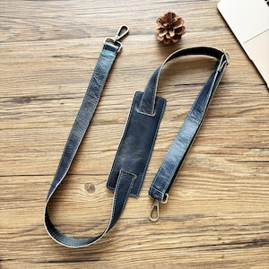 May include: Navy blue leather bag strap with silver-tone metal clasps. The strap includes a padded shoulder rest for comfort. The leather has a textured appearance. The strap is designed to attach to a bag.