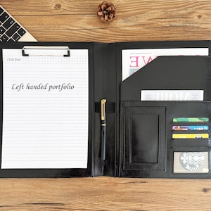 May include: Black leather portfolio with a notepad, pen, and card slots. The notepad is held by a metal clip. The text "Left handed portfolio" is printed on the notepad.