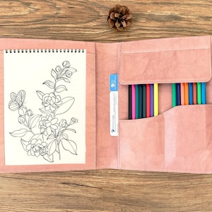 May include: Open pink leather art portfolio with a spiral-bound sketchbook featuring a floral and butterfly line drawing. The portfolio has a pocket with colored pencils and a small blue card. The portfolio is open on a wooden surface.