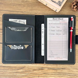 May include: Black leather server book with multiple card slots and a pen holder. A guest check is visible, along with a red pen. The book is open, revealing a section for cash and credit cards.