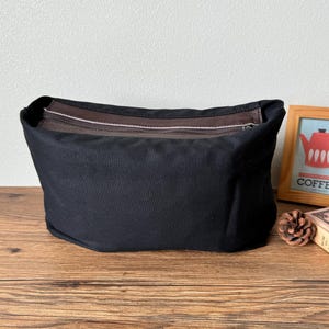 May include: A black canvas zippered pouch with a brown leather trim. The pouch is sitting on a wooden surface.