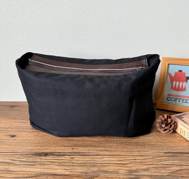 May include: A black canvas zippered pouch with a brown leather trim. The pouch is sitting on a wooden surface.
