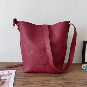 Puede incluir: Un bolso tipo cubo burdeos con un acabado texturizado. El bolso tiene una correa larga y ajustable y otra m&aacute;s corta para llevarlo. El bolso est&aacute; sobre una superficie de madera.