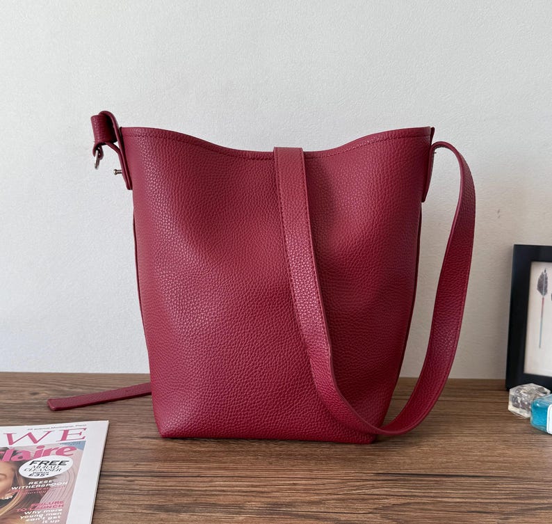 Puede incluir: Un bolso tipo cubo burdeos con un acabado texturizado. El bolso tiene una correa larga y ajustable y otra m&aacute;s corta para llevarlo. El bolso est&aacute; sobre una superficie de madera.
