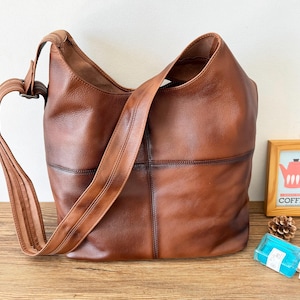 May include: A brown leather shoulder bag with a long strap. The bag has a patchwork design and a large interior. The strap is adjustable and the bag appears to be empty. The bag is sitting on a wooden surface.