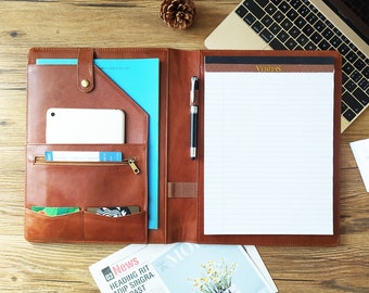 May include: A brown leather portfolio with multiple pockets, a notepad, and a pen. The portfolio is open, revealing a smartphone, cards, and a blue document. A laptop and a newspaper are in the background.