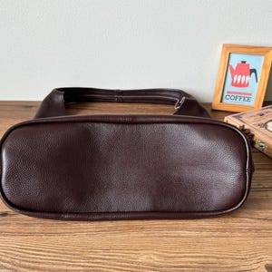 May include: A brown leather handbag with a smooth, textured finish. The bag is shown from the bottom, revealing the stitching and the shape of the bag.
