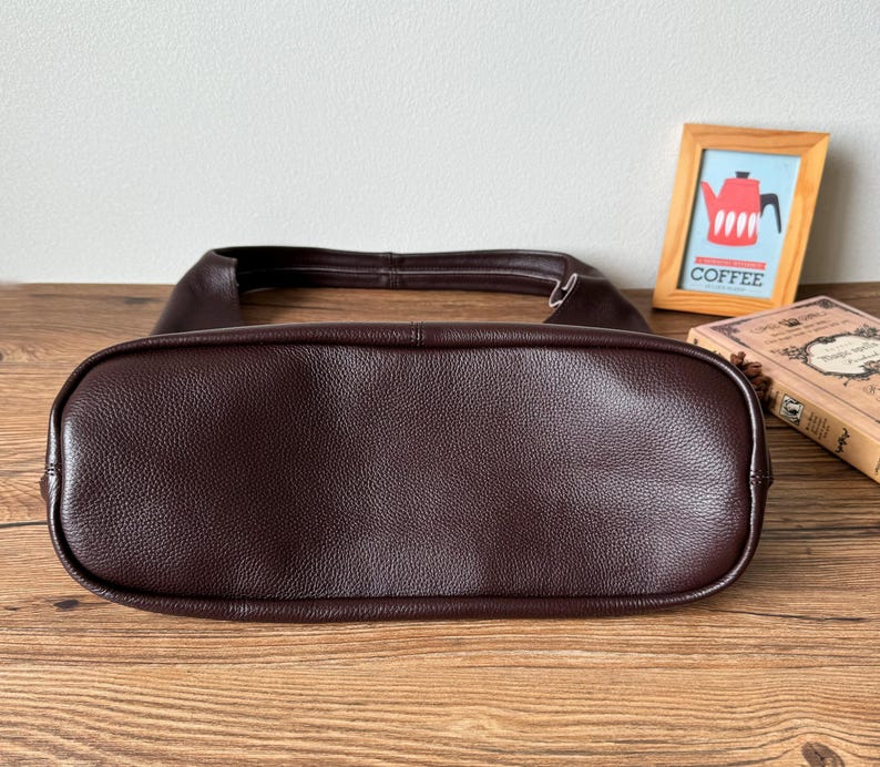 May include: A brown leather handbag with a smooth, textured finish. The bag is shown from the bottom, revealing the stitching and the shape of the bag.