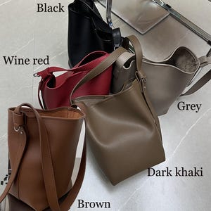 Puede incluir: Una colecci&oacute;n de bolsos tipo cubo de cuero en varios colores: negro, rojo vino, gris, caqui oscuro y marr&oacute;n. Cada bolso tiene una correa para el hombro y un dise&ntilde;o simple y minimalista. Los bolsos se exhiben sobre una superficie clara.
