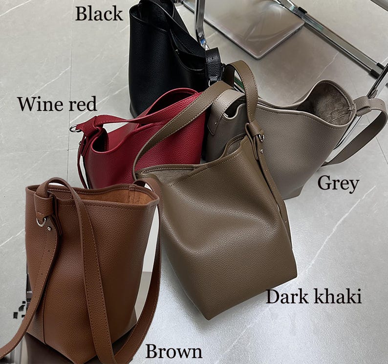 Puede incluir: Una colecci&oacute;n de bolsos tipo cubo de cuero en varios colores: negro, rojo vino, gris, caqui oscuro y marr&oacute;n. Cada bolso tiene una correa para el hombro y un dise&ntilde;o simple y minimalista. Los bolsos se exhiben sobre una superficie clara.