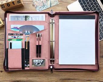 May include: A pink leather binder with a zipper closure, a notepad, and a variety of pockets and slots for pens, cards, and a phone. The binder has a silver ring mechanism and is open to reveal the interior.