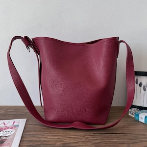 Puede incluir: Un bolso tipo cubo burdeos con una correa larga y ajustable para el hombro. El bolso tiene una superficie texturizada y un dise&ntilde;o sencillo y moderno. El bolso est&aacute; sobre una superficie de madera.