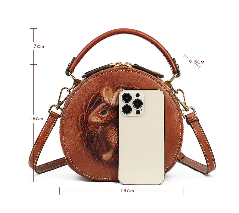 May include: A round, brown leather handbag with a detailed embossed rabbit design. The bag has a top handle and a shoulder strap. It measures 7 inches high, 7 inches deep, and 7 inches wide.
