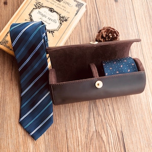 Può includere: Una cravatta blu a righe e una cravatta fantasia sono accanto a un astuccio in pelle marrone per cravatte. La custodia è aperta, rivelando un interno scuro. Un libro vintage con le parole "Magic spells" è sullo sfondo.