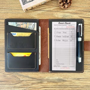 May include: A black leather guest check holder with card slots, a pen, and a guest check. The guest check has handwritten entries for House Salad, Manado Cauliflower, and Chicken Waffles. The holder is open, revealing the interior.