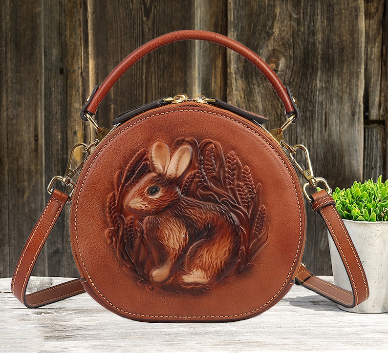 May include: A round, brown leather handbag with a detailed embossed design of a rabbit surrounded by wheat. The bag has a top handle and a detachable shoulder strap. The bag is approximately 8 inches in diameter.
