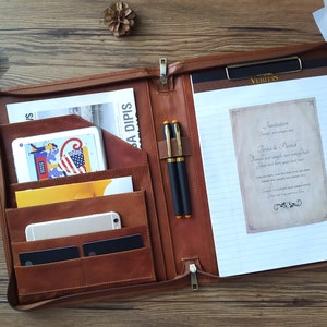 May include: A brown leather portfolio with a zipper closure, multiple pockets, and a built-in notepad. The portfolio is open and has two pens, a tablet, and a phone inside.