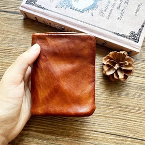 May include: A rectangular, bi-fold wallet crafted from smooth, cognac-colored leather with visible natural grain and tonal variations. The wallet features a simple, unadorned exterior surface with rounded corners and a soft, matte finish.