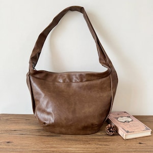 Peut inclure: Un sac à bandoulière en cuir marron avec une longue sangle. Le sac est posé sur une surface en bois à côté d'un livre et d'une pomme de pin. Le sac a une fermeture éclair et une forme arrondie.