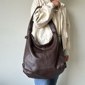 May include: A large, dark brown leather hobo bag with a curved shape. The bag has a single shoulder strap and a zipper closure. The leather has a textured appearance. The bag is being worn over the shoulder.