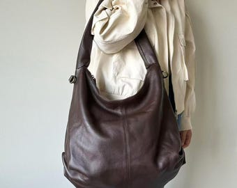 May include: A large, dark brown leather hobo bag with a curved shape. The bag has a single shoulder strap and a zipper closure. The leather has a textured appearance. The bag is being worn over the shoulder.