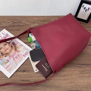 Puede incluir: Un bolso de hombro de color burdeos con una correa larga, abierto para revelar su contenido. En el interior hay un smartphone, una tableta, un pasaporte y varios frascos peque&ntilde;os. Una revista con el texto "Spring is on" est&aacute; cerca.