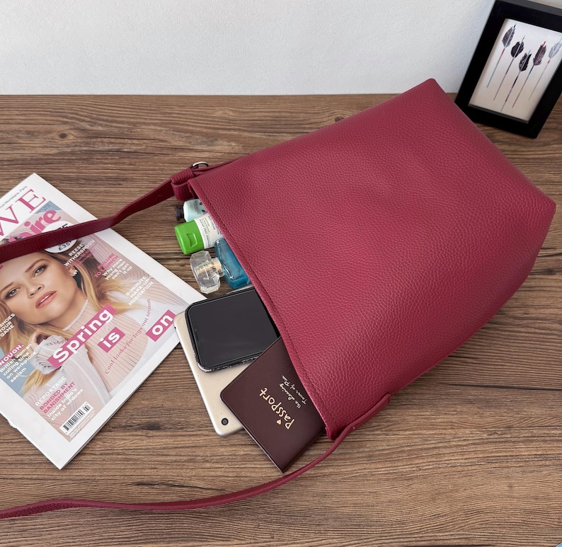 Puede incluir: Un bolso de hombro de color burdeos con una correa larga, abierto para revelar su contenido. En el interior hay un smartphone, una tableta, un pasaporte y varios frascos peque&ntilde;os. Una revista con el texto "Spring is on" est&aacute; cerca.