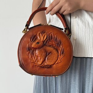 May include: A round, brown leather handbag with a detailed embossed design of a rabbit surrounded by wheat. The bag has a top handle and a zipper closure. The bag is held by a person.