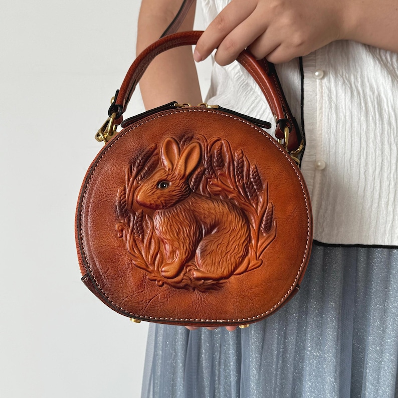 May include: A round, brown leather handbag with a detailed embossed design of a rabbit surrounded by wheat. The bag has a top handle and a zipper closure. The bag is held by a person.