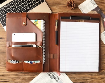 May include: A brown leather portfolio with a notepad, a pen, and a tablet. The portfolio has a zippered pocket, a flap pocket, and two card slots. The notepad has a spiral binding and is open to a blank page. The pen is black with a silver clip. The tablet is white and has a black screen. The portfolio is on a wooden surface.