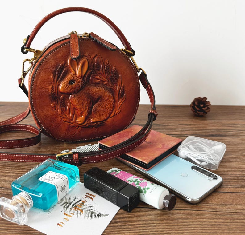 May include: A brown leather round handbag with a carved rabbit design. The bag has a top handle and a shoulder strap. Other items include a phone, perfume, and cosmetics on a wooden surface.
