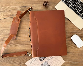 May include: A brown leather portfolio with a long adjustable strap. The portfolio has a flap closure and a metal clasp. The portfolio is lying on a wooden surface.