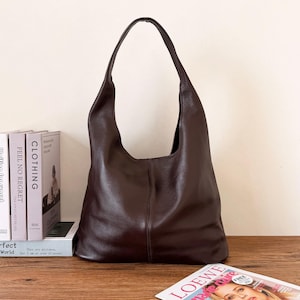May include: A dark brown leather hobo bag with a soft, slouchy silhouette and a single shoulder strap. The bag features a vertical seam down the center of the front panel, a pebbled texture, and a spacious main compartment with a curved top opening.