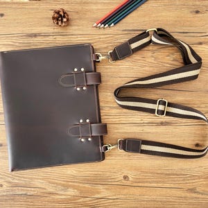 May include: A dark brown leather portfolio with two buckle closures and a detachable striped shoulder strap. The portfolio has metal hardware and is designed for carrying documents or a laptop.