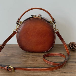 May include: A round, brown leather handbag with a top handle and a detachable shoulder strap. The bag features a zipper closure and gold-colored hardware. The stitching is white, and a small pinecone sits on the wooden surface.